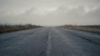 Empty asphalt road stretching into distance under overcast sky in rural countryside landscape with grass fields on both sides creating moody atmosphere.
