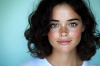 Young woman with natural freckles, dark curly hair and blue eyes smiling at camera against turquoise background, radiating genuine happiness and confidence.