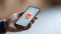 Mobile phone displaying orange shopping cart icon on white screen, held in hand against blurred background. Perfect for e-commerce and mobile shopping concepts.