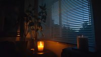 Moody evening ambiance with glowing candles on windowsill, venetian blinds casting shadows, and silhouette of potted plant creating cozy atmospheric scene.