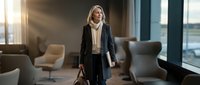 Professional businesswoman in elegant black coat walking through modern office lobby with leather handbag during golden hour sunlight streaming through windows.
