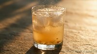 Chilled amber cocktail with ice cubes in a rocks glass, sparkling bubbles rising, warm golden sunlight casting long shadows on stone surface.