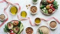 Overhead view of organic vegetables, nuts, seeds, and olive oil arranged on white surface with pink ribbons, showcasing healthy eating and nutritious ingredients for cooking.