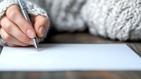 Female hand with natural manicure holding silver pen over blank white paper, wearing cozy knit sweater, ready for writing or creative work.