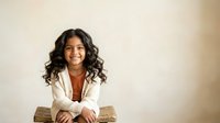 Young woman with long dark wavy hair and medium brown skin tone, wearing a white blazer over a rust-colored top, seated and smiling at camera against neutral beige background for corporate and professional use.