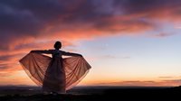 Young woman silhouette dancing with flowing fabric against dramatic sunset sky. Freedom and movement concept for wellness and lifestyle content.