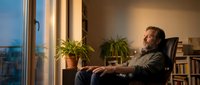 Middle-aged man with light skin and reddish-brown hair sitting in black office chair by large window at sunset, relaxing in modern home office with potted plants and bookshelves, contemplative mood.