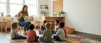 Female teacher with long brown hair reading a storybook to a diverse group of young children sitting on a colorful rug in a bright classroom with wooden furniture and plants.