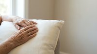 Elderly woman's hands resting on white pillow in bright bedroom. Senior care, comfort, rest, peaceful moments at home for healthcare and wellness concepts.