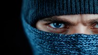 Close-up view of intense blue eyes peering through dark knitted balaclava mask against black background, creating mysterious and dramatic atmosphere.