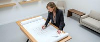 Professional businesswoman architect reviewing construction blueprints at modern office desk. Corporate planning meeting workspace interior design.
