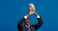 Successful African American male professional in dark business suit and sunglasses holding trophy overhead against blue background, celebrating achievement and victory.