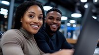 Young African American professionals working together at laptop computer, smiling and collaborating in modern office environment with blurred background.