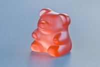 Red translucent gummy bear candy sitting on a light blue-gray surface, soft studio lighting highlighting its glossy texture and cheerful shape.