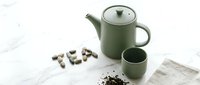 Sage green ceramic teapot and cup set on marble surface with loose leaf tea and pebbles, minimal Japandi style kitchen decor.