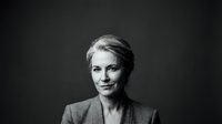 Mature woman with blonde hair styled back, fair complexion, wearing professional blazer against dark background, studio portrait photography.
