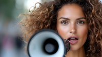 Young mixed race woman with curly hair holding megaphone, close up portrait showing confident expression and natural makeup against blurred background.