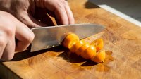 Caucasian male hand slicing fresh carrot on wooden cutting board with chef knife in bright kitchen lighting for cooking and food preparation.