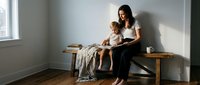Young mother and toddler daughter reading book together on wooden bench by window in minimalist home interior with natural lighting for family bonding.