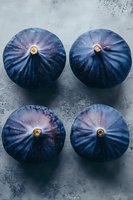 Four fresh purple figs arranged on textured stone surface, top view of ripe Mediterranean fruit with natural ridged pattern for culinary and healthy eating concepts.