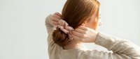 Young woman with reddish-brown hair wearing a beige knit sweater, styling her hair into a bun with a pink fabric scrunchie against a white background.