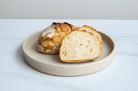 Fresh crusty artisan bread loaf sliced on minimal ceramic plate against white marble background, showing soft interior crumb texture and golden crust.