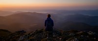 Hiker in dark jacket standing on mountain peak at sunset, overlooking layered valleys and distant hills in golden hour light. Adventure and outdoor exploration.