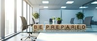 Young professional woman with light skin tone in business casual attire sitting at modern office desk with wooden blocks spelling "BE PREPARED" for corporate training, motivation, readiness and workplace preparation concepts.