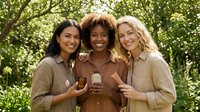 Three diverse women friends enjoying coffee together outdoors in natural sunlight, smiling and bonding in casual earth tone clothing among green foliage.