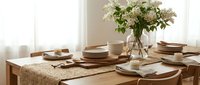 Elegant dining table set with neutral tableware, woven placemat, and fresh white flowers in glass vase creating warm, inviting atmosphere for home dining.