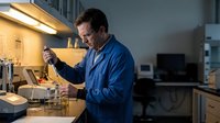 Middle-aged Caucasian male scientist in blue lab coat conducting research experiment in modern laboratory with test tubes and equipment under bright lighting.