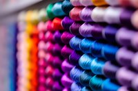 Colorful rows of thread spools in vibrant shades arranged diagonally, with selective focus creating artistic blur effect. Macro shot showing textile crafting supplies.