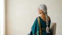 Senior woman with long gray braided hair wearing teal cardigan and dark top, sitting on chair facing away, looking toward window light for wellness and lifestyle content.