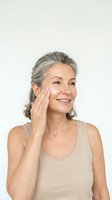 Mature woman applying face cream with gentle touch, showcasing anti-aging skincare routine and natural beauty confidence on neutral background.