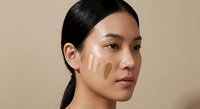 Young Asian woman applying foundation makeup with fingers on face against neutral beige background for skincare and cosmetics advertising campaigns.