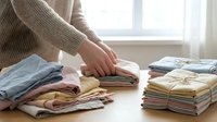 Hands organizing and folding stacked colorful clothing and textiles on wooden table near bright window, laundry and home organization concept.