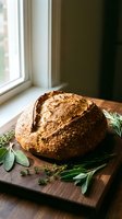 Rustic artisan sourdough bread loaf on wooden cutting board with fresh herbs by window light for bakery and homemade cooking concepts.