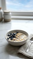 Creamy oatmeal bowl topped with fresh blueberries, sliced almonds, and seeds served on marble countertop by bright window for breakfast and healthy eating content.