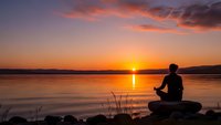 Man meditating by tranquil lake at sunset with orange sky reflection on water creating peaceful zen atmosphere for wellness and mindfulness concepts.