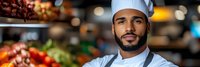 Professional male chef with beard wearing white uniform and hat standing confidently in modern restaurant kitchen with fresh ingredients and cooking equipment.