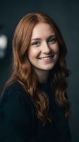 Young Caucasian woman with long wavy red hair smiling warmly at camera wearing dark sweater against blurred background for professional headshot photography.