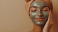 Young African American woman with clay face mask and orange towel wrapped around head enjoying spa skincare treatment against neutral beige background.