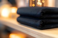 Folded black towels on wooden shelf with soft bokeh lights in background creating spa-like atmosphere for wellness and relaxation concepts.