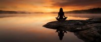 Woman in lotus position meditating on rocky outcrop at sunrise, silhouetted against golden sky with water reflection below for wellness and mindfulness concepts.