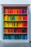Books arranged by color spectrum on white wooden bookshelf creating rainbow pattern. Decorative library organization displays vibrant spines from yellow to red and blue.