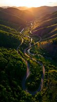 Winding mountain road through lush green valley with river at golden hour sunrise, aerial view of serpentine highway cutting through forested hills and landscape.