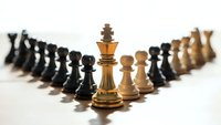 Golden chess king piece standing prominently in front of arranged black and white chess pieces on light background for strategic business concepts.