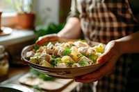 Fresh potato salad with herbs and vegetables served in rustic ceramic bowl, hands in plaid shirt holding dish against window light, cozy home cooking atmosphere.