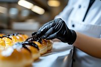 Professional pastry chef in black gloves decorating fresh baked eclairs with chocolate sauce in commercial kitchen, close up view of confectionery preparation process.