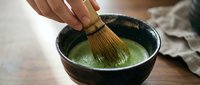 Matcha tea preparation with bamboo whisk stirring powdered green tea in black ceramic bowl on wooden surface.